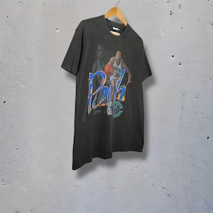 May include: A black t-shirt with a basketball player graphic and the name "RB" in blue and green. The shirt has a vintage appearance and is suspended from a wooden hanger.