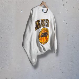 Könnte beinhalten: Cremefarbenes Sweatshirt mit dem Text "LONG BEACH STATE" in gold-schwarzen Buchstaben. Ein kreisförmiges Emblem zeigt die Worte "CALIFORNIA STATE UNIV" und "49ERS" in einem Sonnenstrahl-Design. Das Sweatshirt hängt an einem Holzkleiderbügel.