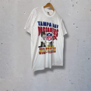 Könnte beinhalten: Weißes T-Shirt mit dem Logo der Tampa Bay Buccaneers und einem Cartoon-Footballspieler. Das Shirt zeigt "TAMPA BAY BUCCANEERS" in Rot und Blau sowie "WE WON'T BE VIC-TIMED" in Gelb und Rot.