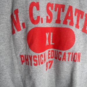 K&ouml;nnte beinhalten: Ein graues Sweatshirt mit roter Schrift. Der Text lautet "N.C. STATE" &uuml;ber einem roten Oval mit "XL" darin. Darunter steht "PHYSICAL EDUCATION 97". Das Sweatshirt hat einen V-Ausschnitt.