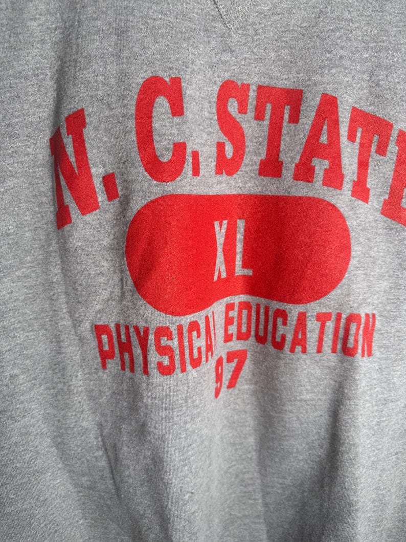 K&ouml;nnte beinhalten: Ein graues Sweatshirt mit roter Schrift. Der Text lautet "N.C. STATE" &uuml;ber einem roten Oval mit "XL" darin. Darunter steht "PHYSICAL EDUCATION 97". Das Sweatshirt hat einen V-Ausschnitt.