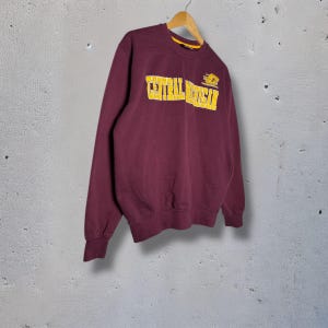 May include: A maroon sweatshirt with the text "CENTRAL MICHIGAN" in yellow block letters. The sweatshirt is hanging on a wooden hanger against a gray background. A small yellow logo is to the right of the text.