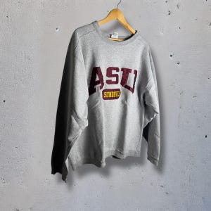Peut inclure: Un sweat-shirt gris avec les lettres "ASU" bordeaux et or. Le mot "SUNDEVILS" est imprimé en or en dessous. Le sweat-shirt est suspendu à un cintre en bois.