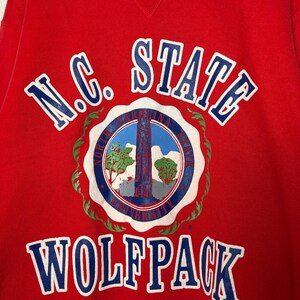 Op de afbeelding: Een rode sweatshirt met de woorden "N.C. STATE WOLF PACK" in witte en blauwe letters. De sweatshirt heeft een cirkelvormig embleem met een blauw-wit ontwerp, inclusief een toren en bomen.