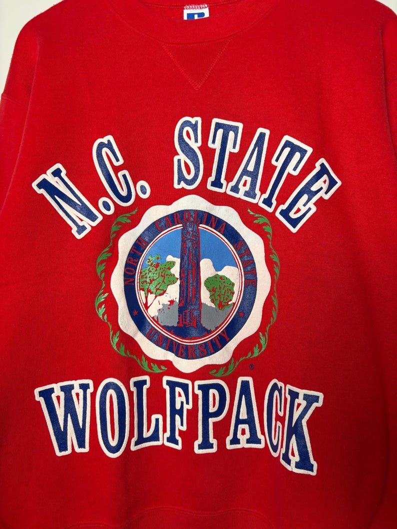 Op de afbeelding: Een rode sweatshirt met de woorden "N.C. STATE WOLF PACK" in witte en blauwe letters. De sweatshirt heeft een cirkelvormig embleem met een blauw-wit ontwerp, inclusief een toren en bomen.