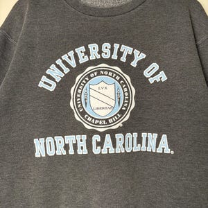 Op de afbeelding: Donkergrijze sweatshirt met de tekst "UNIVERSITY OF NORTH CAROLINA" in lichtblauwe en witte letters. Het universiteitszegel staat in het midden. De sweatshirt hangt aan een houten kleerhanger.
