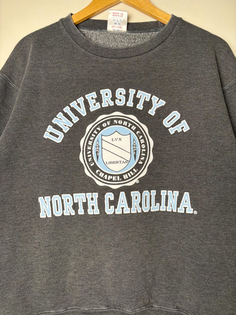 Op de afbeelding: Donkergrijze sweatshirt met de tekst "UNIVERSITY OF NORTH CAROLINA" in lichtblauwe en witte letters. Het universiteitszegel staat in het midden. De sweatshirt hangt aan een houten kleerhanger.
