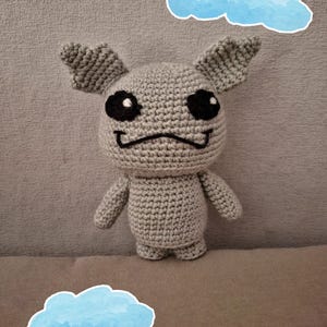 Puede incluir: Una criatura gris hecha a mano con ganchillo, con grandes ojos negros, brazos pequeños y orejas. La criatura tiene una boca cosida y está de pie frente a un fondo gris con dos pegatinas de nubes azules de acuarela.