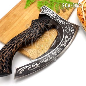 May include: A decorative axe with a dark wooden handle and a silver blade. The blade features intricate engraved designs and the text "SCA-0027". The axe is displayed on a wooden cutting board.