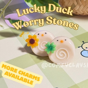 May include: Two Lucky Duck Worry Stones, each with a white base, a spiral design, and a small orange beak. One has a yellow sunflower charm, the other a green clover. The text "Lucky Duck Worry Stones" is displayed.