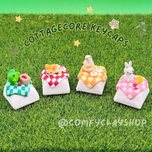 May include: Four cottagecore-themed keycaps on a green surface. Each keycap features a different design: a frog with a basket of berries, a dog, a bird with flowers, and a bunny. The keycaps are on white bases with checkered patterns.