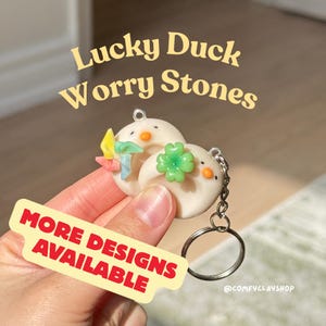 May include: Two white worry stones shaped like ducks, each with a small orange beak and black eyes. One has a green four-leaf clover, the other a colorful pinwheel. The text "Lucky Duck Worry Stones" is above the ducks, and "More Designs Available" is on a yellow banner.