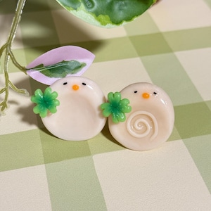 May include: Two round, off-white decorative objects with black eyes and orange beaks, each holding a green four-leaf clover. One has a spiral design. The objects are on a green and white checkered surface.
