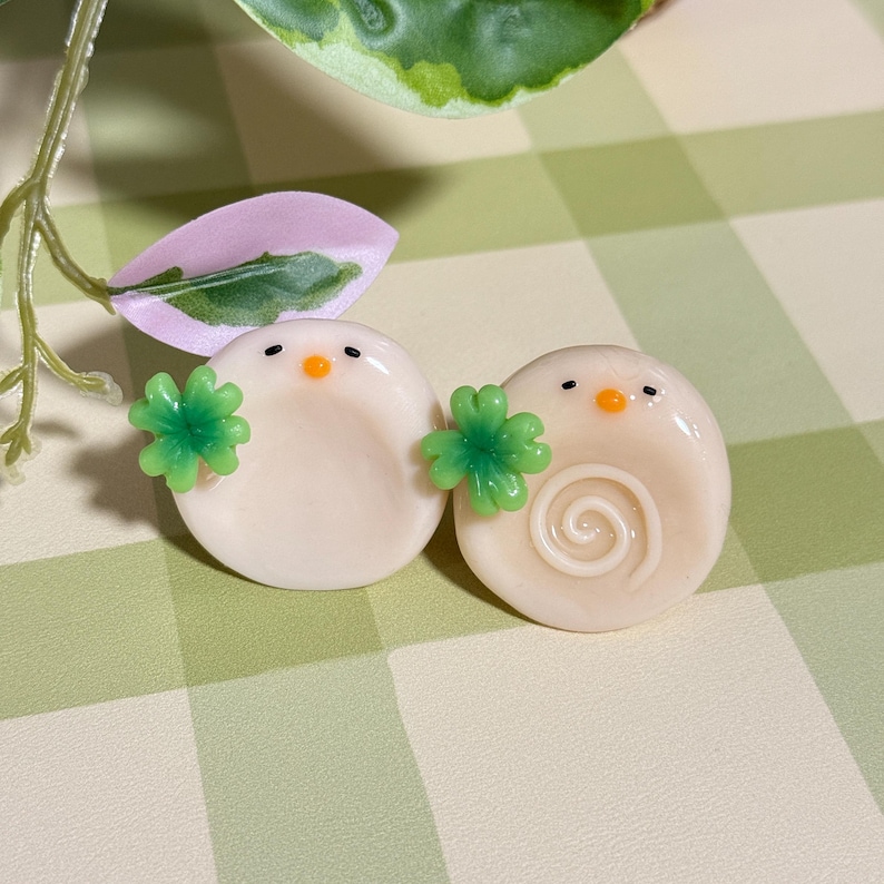 May include: Two round, off-white decorative objects with black eyes and orange beaks, each holding a green four-leaf clover. One has a spiral design. The objects are on a green and white checkered surface.