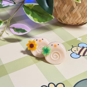 May include: Two cream-colored, snail-shaped earrings with orange beaks and black eyes. One earring has a yellow sunflower, the other a green flower. The earrings are on a green and white checkered surface.