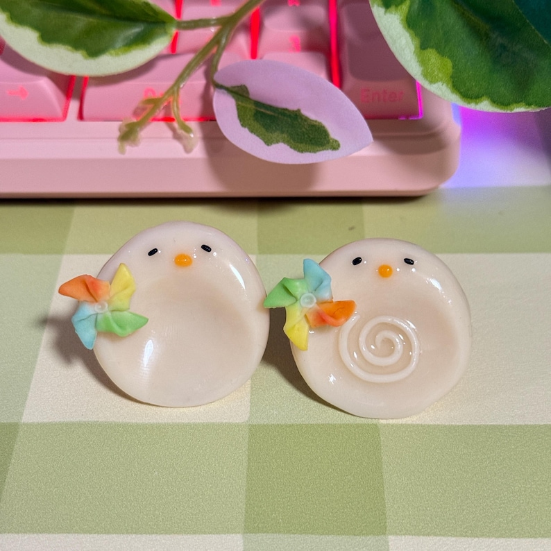 May include: Two off-white, egg-shaped figurines with black dot eyes and orange beaks. Each has a colorful pinwheel attached. One has a swirl design. The figurines are on a green and white checkered surface.