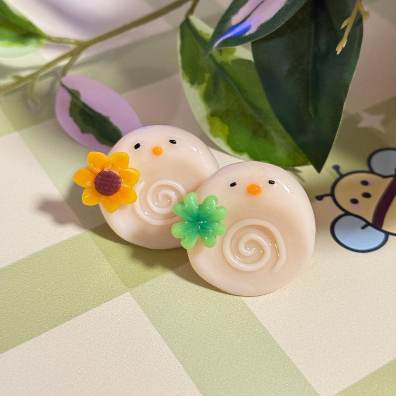 May include: Two off-white, snail-shaped decorative objects with spiral designs. Each has a small orange beak and black eyes. One has a yellow and brown sunflower, the other a green flower. The background is a light green and white checkered pattern.