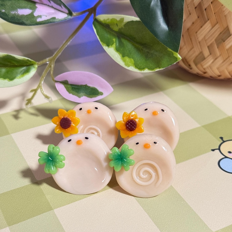 May include: Four small, round, off-white decorative objects with a bird-like design. Two have yellow sunflower accents, and two have green clover accents. Each has a small orange beak and black eyes. The objects are on a green and white checkered surface.
