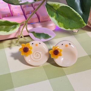 May include: Two off-white, egg-shaped decorative objects with small black eyes and orange beaks. Each has a yellow sunflower attached. One has a spiral design. The background includes a pink keyboard and green and white leaves.