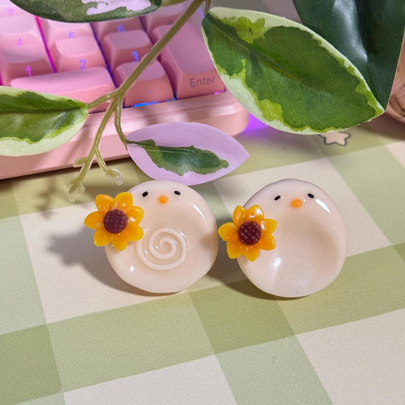 May include: Two off-white, egg-shaped decorative objects with small black eyes and orange beaks. Each has a yellow sunflower attached. One has a spiral design. The background includes a pink keyboard and green and white leaves.