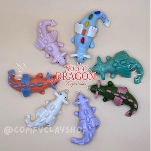 Puede incluir: Seis llaveros con forma de dragón en varios colores, incluyendo azul claro, rosa, naranja, verde azulado, verde y morado, dispuestos en círculo. El texto JELLY DRAGON Keychains y @COMFYCLAYSHOP está impreso en el centro.
