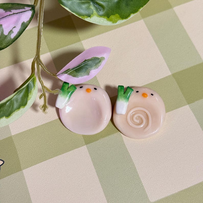 May include: Two light pink, round, decorative objects with green and white accents, resembling cartoon characters. One has a spiral design. The objects have small black eyes and orange beaks. The background is a green and white checkered pattern.