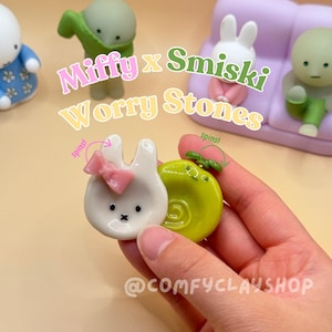 Könnte beinhalten: Eine Sammlung von Miffy x Smiski Worry Stones. Das Bild zeigt ein weißes Kaninchen mit einer rosa Schleife, eine grüne Schnecke und andere Figuren. Der Text "Miffy x Smiski Worry Stones" ist im Bild zu sehen.