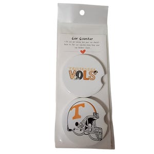 May include: Two white car coasters with a Tennessee Vols logo and a football helmet design. The coasters are packaged in a clear plastic sleeve with a white card that reads "Car Coaster" and "Life can get messy, but your car doesn't have to. Our car coasters help keep your cup holders clean."