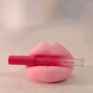 May include: A bright pink lip-shaped object holding a red lip product. The lip product has a red cap and a clear tube. The text "peripera" is printed on the lip product. The background is a soft, blurred pink.