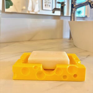 May include: A yellow soap dish shaped like a block of Swiss cheese with a white bar of soap inside.