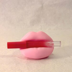 May include: A pink lip-shaped holder with a red lip gloss. The lip gloss has the word "peripera" printed on it. The holder and lip gloss are set against a light background.