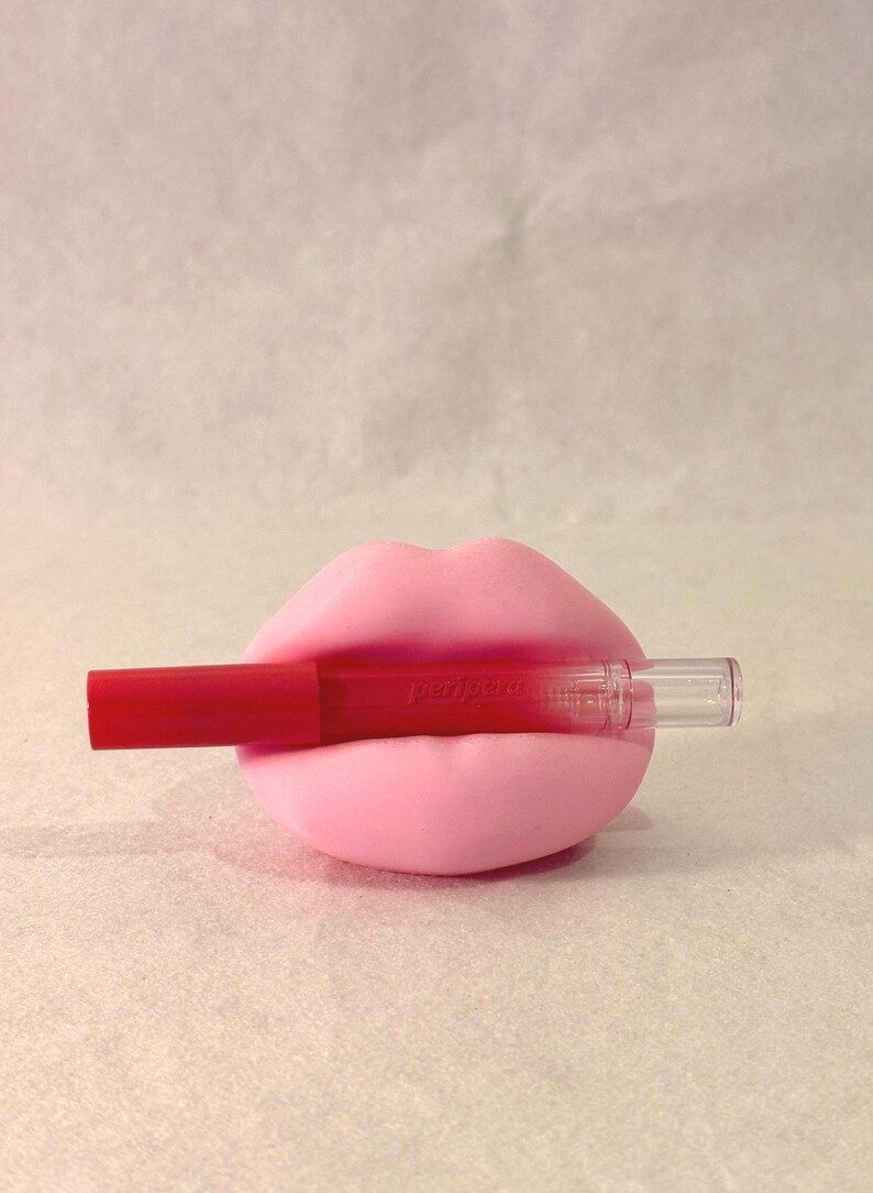May include: A pink lip-shaped holder with a red lip gloss. The lip gloss has the word "peripera" printed on it. The holder and lip gloss are set against a light background.
