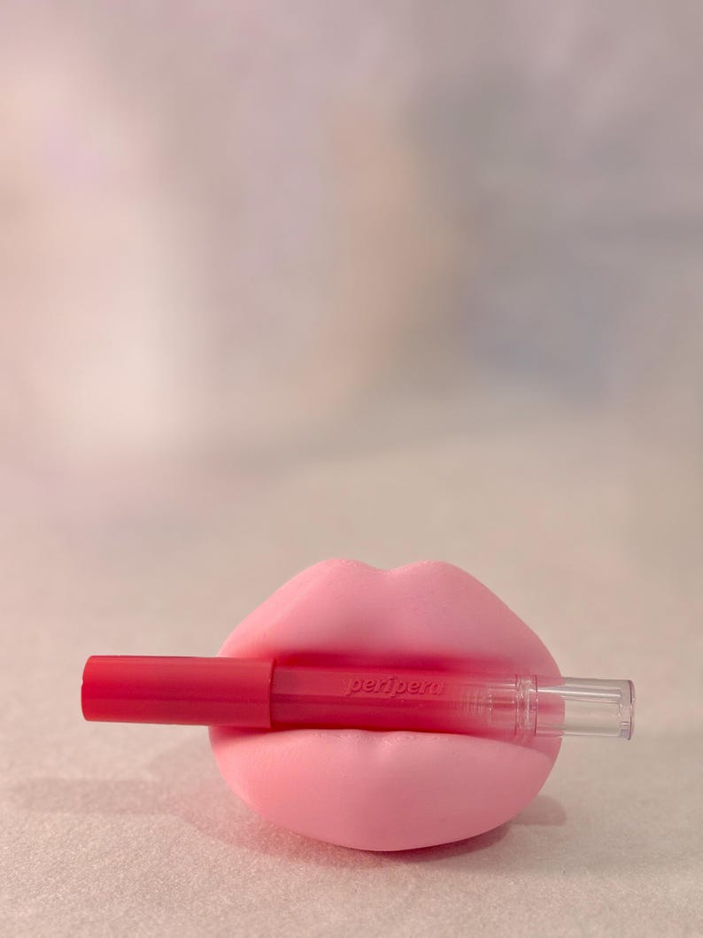 May include: A bright pink lip-shaped object holding a red lip product. The lip product has a red cap and a clear tube. The text "peripera" is printed on the lip product. The background is a soft, blurred pink.