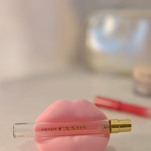 May include: A pink lip-shaped perfume holder cradles a small Prada Candy perfume bottle. The bottle is clear with gold accents and the words "PRADA CANDY" are printed on the bottle. The background is blurred.