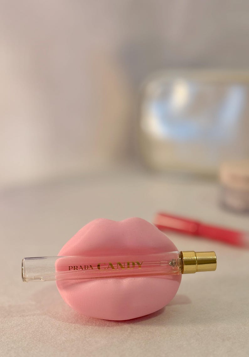 May include: A pink lip-shaped perfume holder cradles a small Prada Candy perfume bottle. The bottle is clear with gold accents and the words "PRADA CANDY" are printed on the bottle. The background is blurred.