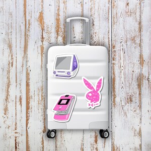 Puede incluir: Una maleta blanca con tres pegatinas: un ordenador morado y blanco, un tel&eacute;fono plegable rosa y un logotipo de conejo rosa brillante. Las pegatinas est&aacute;n adheridas a la parte delantera de la maleta.