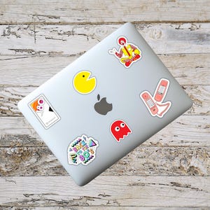 May include: A collection of colorful stickers on a laptop. Stickers include a yellow Pac-Man, a red ghost, a pink telephone, a Ronald McDonald figure, and a sticker with the text "Back to the 80's". The laptop is silver.