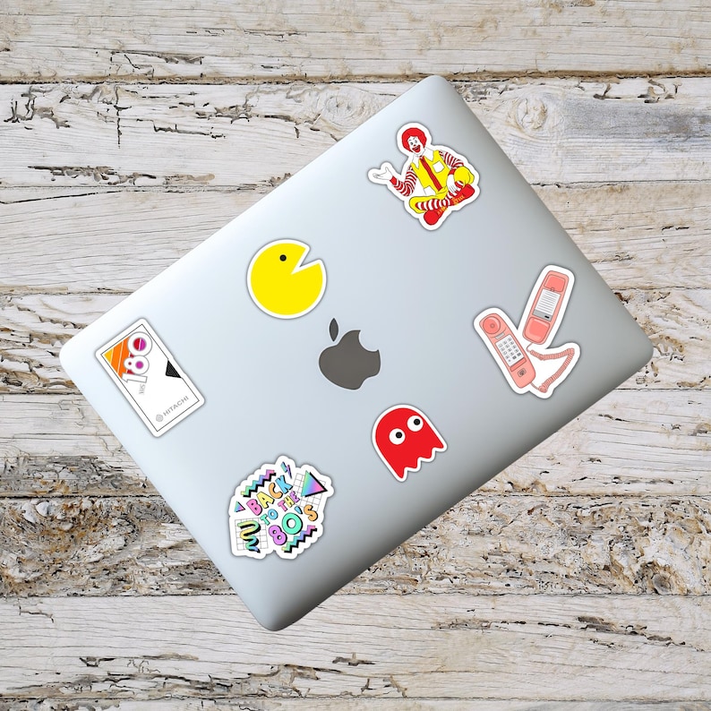 May include: A collection of colorful stickers on a laptop. Stickers include a yellow Pac-Man, a red ghost, a pink telephone, a Ronald McDonald figure, and a sticker with the text "Back to the 80's". The laptop is silver.