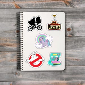 May include: A spiral-bound notebook featuring a collection of colorful stickers. The stickers include a silhouette of a person on a bicycle, a pizza parlor sign, a unicorn, a Ghostbusters logo, and a vintage instant camera.