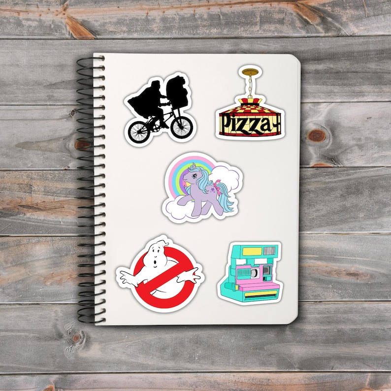 May include: A spiral-bound notebook featuring a collection of colorful stickers. The stickers include a silhouette of a person on a bicycle, a pizza parlor sign, a unicorn, a Ghostbusters logo, and a vintage instant camera.