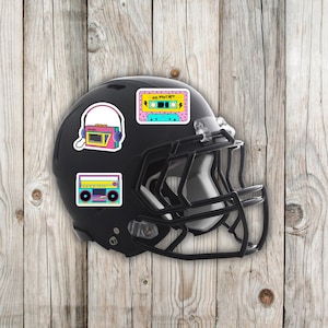 May include: A black football helmet adorned with colorful stickers featuring retro cassette tapes, boomboxes, and a radio with headphones. The stickers have a vibrant 80s aesthetic, with bright pink, yellow, and teal accents.