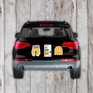 May include: Black SUV with three retro-themed stickers: a yellow 1980s design, a Polaroid HS sticker with a rainbow stripe, and a yellow Pac-Man ghost. The stickers are on the rear of the vehicle.