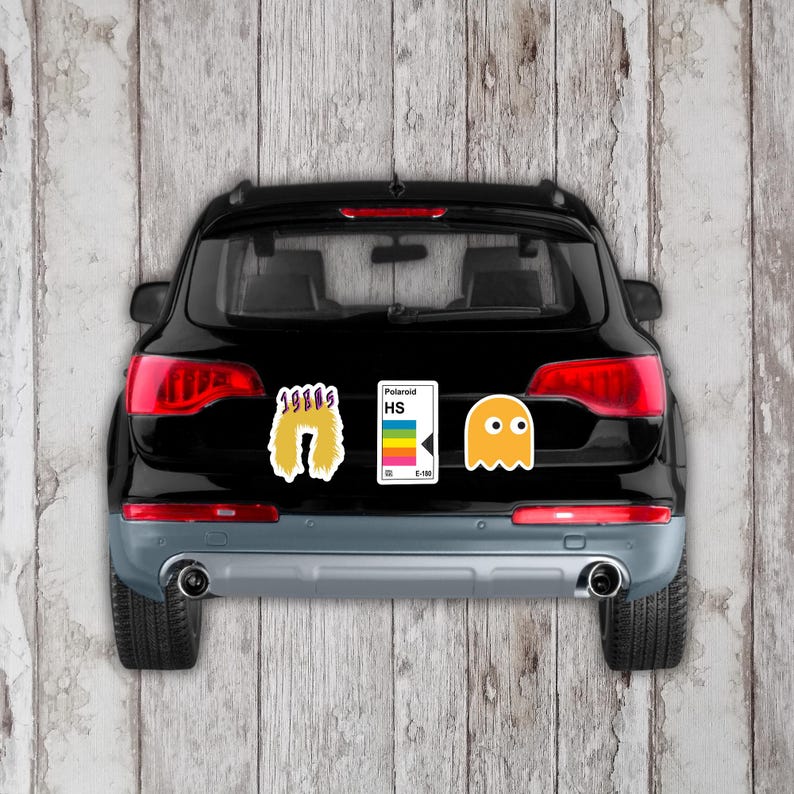 May include: Black SUV with three retro-themed stickers: a yellow 1980s design, a Polaroid HS sticker with a rainbow stripe, and a yellow Pac-Man ghost. The stickers are on the rear of the vehicle.