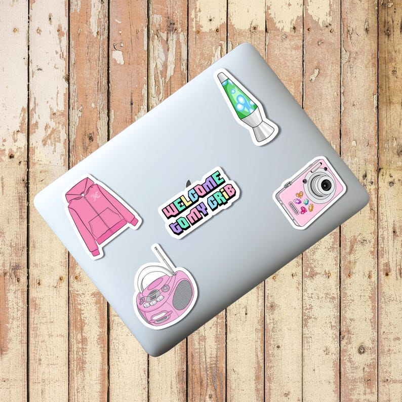 Puede incluir: Un port&aacute;til con pegatinas: una sudadera con capucha rosa, una l&aacute;mpara de lava, una c&aacute;mara rosa, una radio rosa y una pegatina con el texto "WELCOME TO MY CRIB" en colores arco&iacute;ris. El port&aacute;til est&aacute; sobre una superficie de madera.