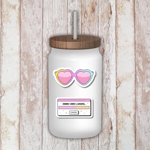 Puede incluir: Tarro de vidrio blanco con tapa y pajita de madera. El tarro tiene pegatinas de gafas de sol en forma de coraz&oacute;n con degradados rosa, amarillo y azul, y una pegatina con el texto "2000s Vibes Loading..." y una barra de progreso.