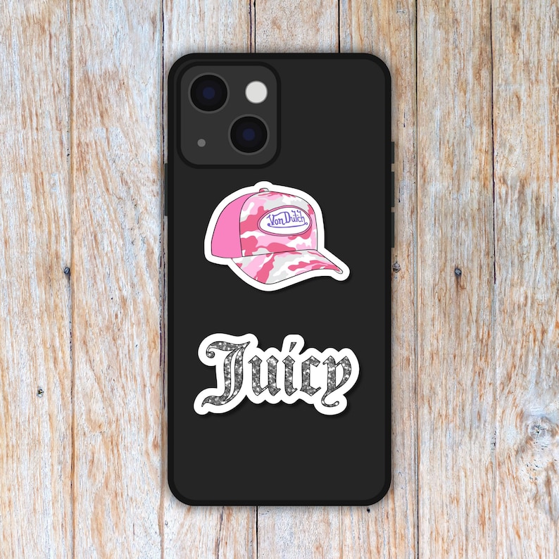 Puede incluir: Una funda de tel&eacute;fono negra con dos pegatinas. La pegatina superior es una gorra de b&eacute;isbol rosa y blanca de camuflaje con el texto "Von Dutch". La pegatina inferior dice "Juicy" en una fuente brillante.