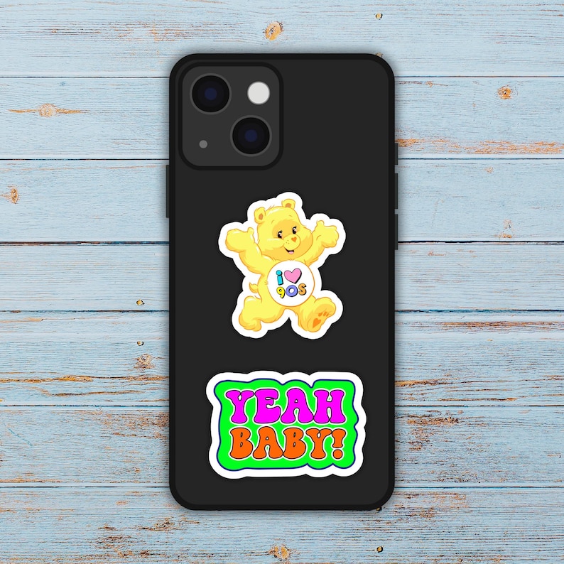 May include: Black phone case with two stickers. The top sticker is a yellow bear with the text "I 90s" in a heart. The bottom sticker says "YEAH BABY!" in pink and orange.