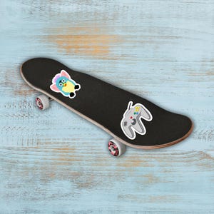 May include: A black skateboard with a Furby sticker and a gray video game controller sticker. The skateboard has pink and white wheels and is set against a light blue wooden background.