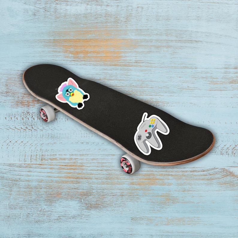 May include: A black skateboard with a Furby sticker and a gray video game controller sticker. The skateboard has pink and white wheels and is set against a light blue wooden background.