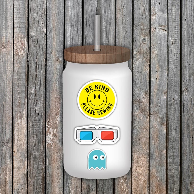 May include: White glass tumbler with a wooden lid and straw. The tumbler is decorated with stickers, including a yellow smiley face with the text "BE KIND PLEASE REWIND", 3D glasses, and a blue ghost.